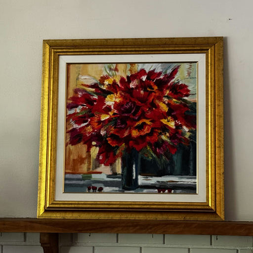 A vibrant painting of a red flower bouquet in a dark vase, framed in gold, hangs on a wall above a wooden shelf. The background features soft hues.
