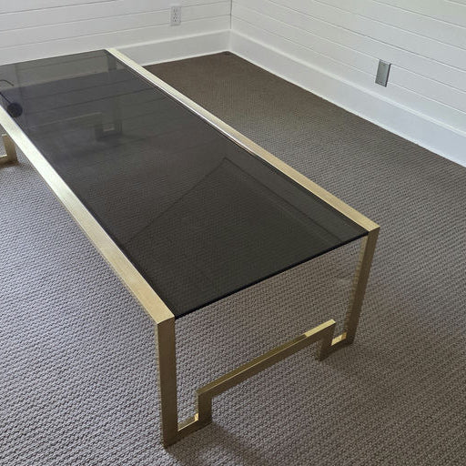 A modern coffee table with a sleek black glass top and gold metal frame sits on a neutral, textured carpet in a sparsely furnished room.