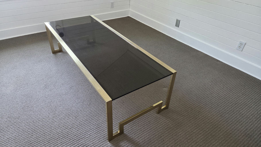 A modern coffee table with a sleek black glass top and gold metal frame sits on a neutral, textured carpet in a sparsely furnished room.