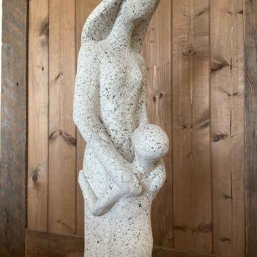 A stylized stone sculpture of a woman holding a child close, conveying themes of nurturing and motherhood, set against a wooden background.