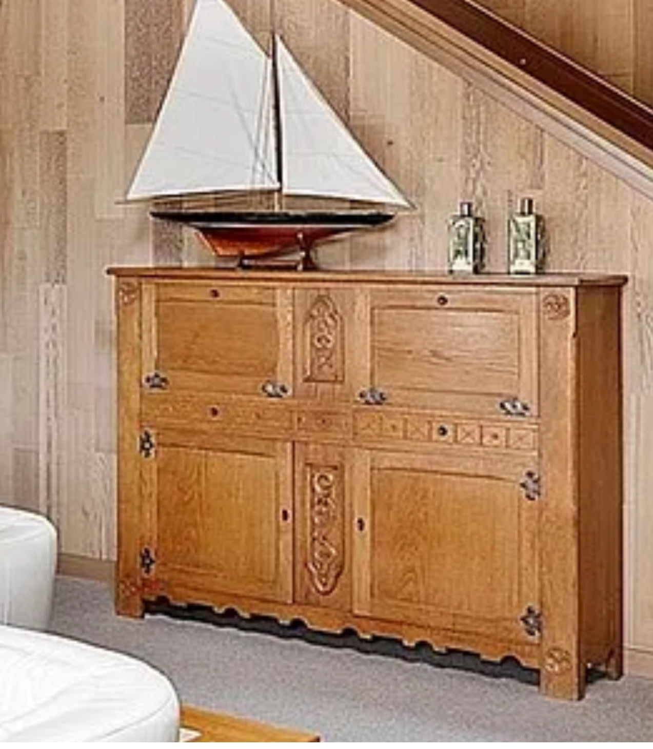 Wooden cabinet with decorative carvings, featuring a model sailboat on top and two bottles beside it, set against a wooden wall.