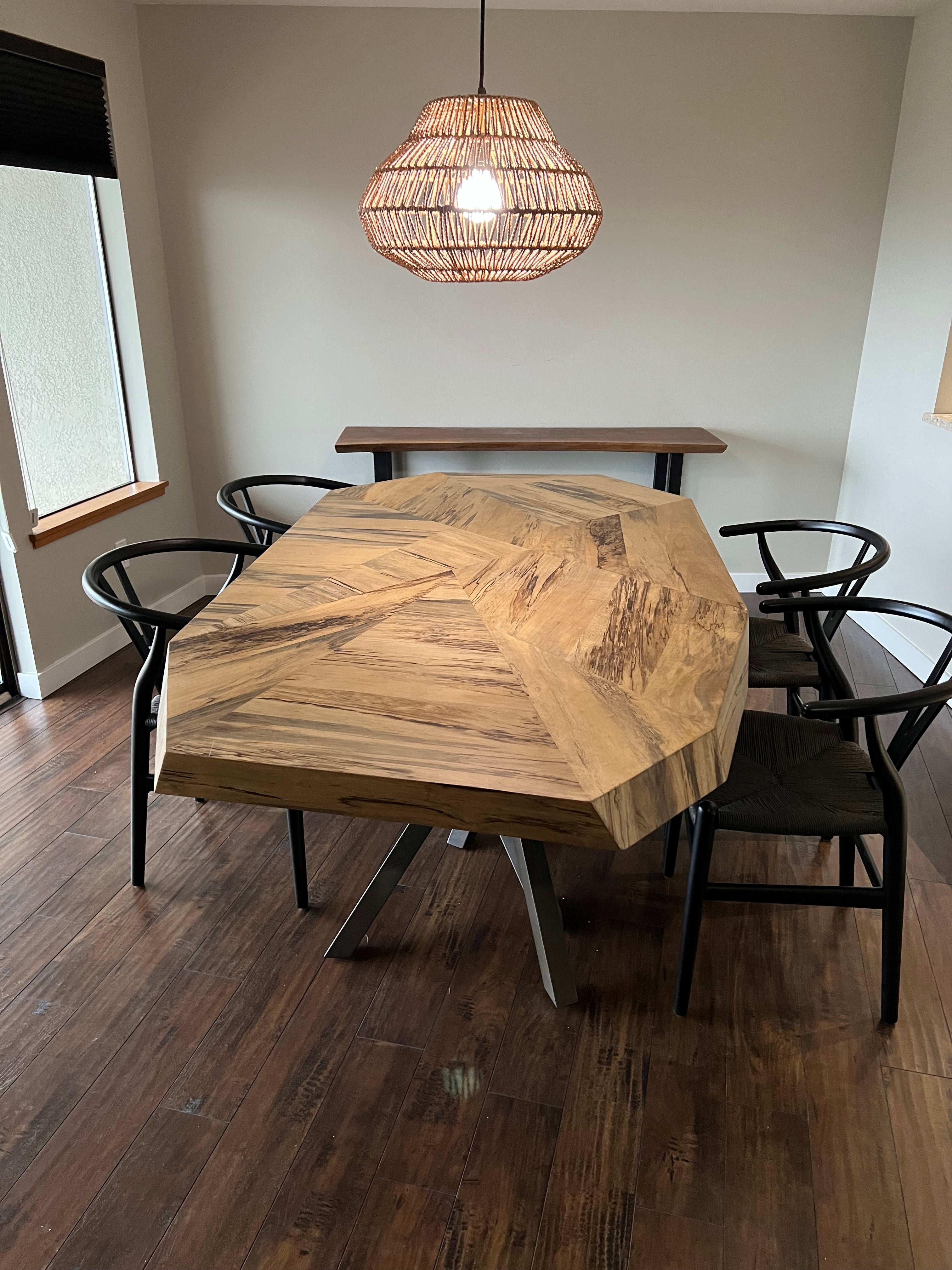 A modern dining area featuring a unique octagonal wooden table, four black chairs, and a woven pendant light, all set on dark wooden flooring.
