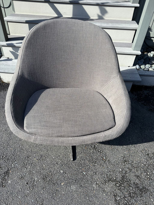 A gray, upholstered chair with a rounded back and cushioned seat sits on a gravel surface, in front of wooden steps and a pale wall.