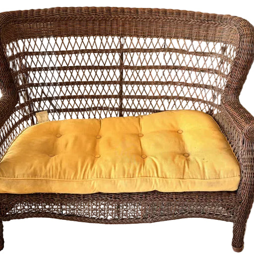 A wicker loveseat with a bright yellow cushion sits on carpet, near a dining table. A red backpack and a green bag are visible in the background.