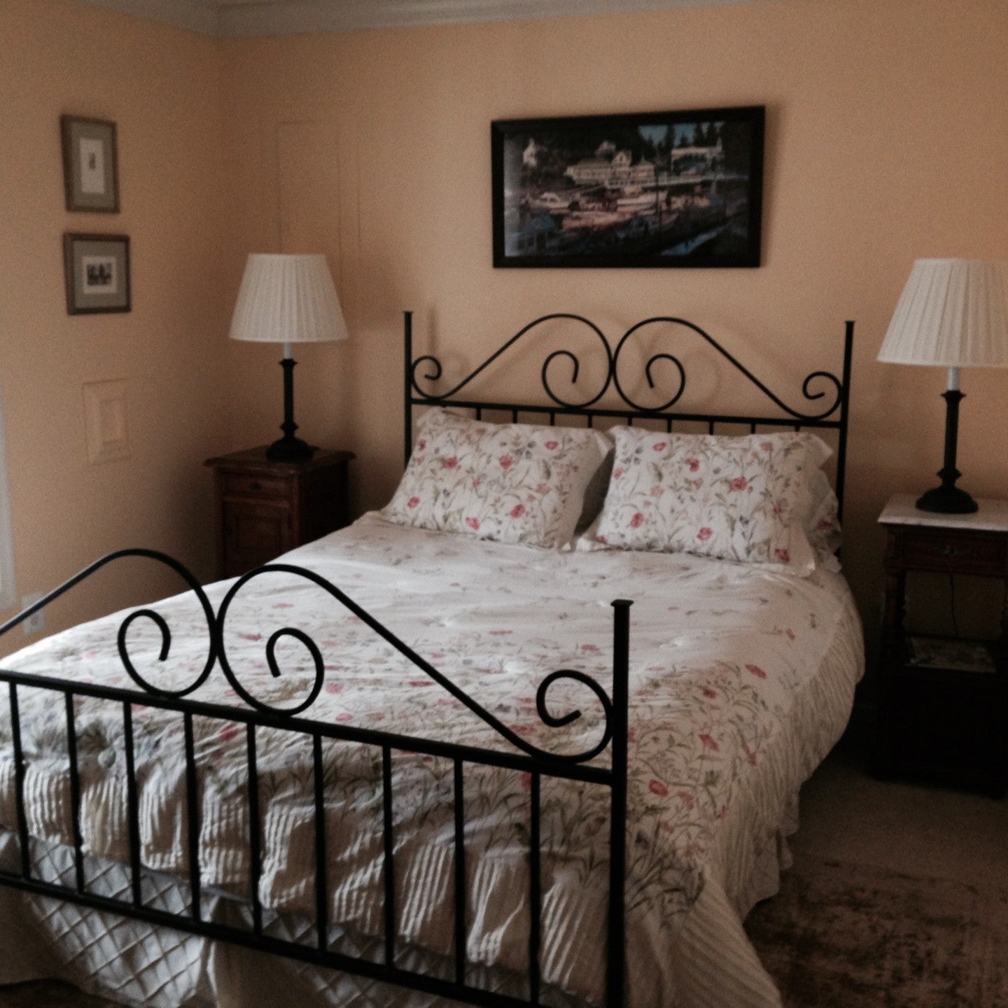 A cozy bedroom with a black iron bed, floral bedding, two bedside lamps, and a painting on the wall, illuminated by natural light from a window.