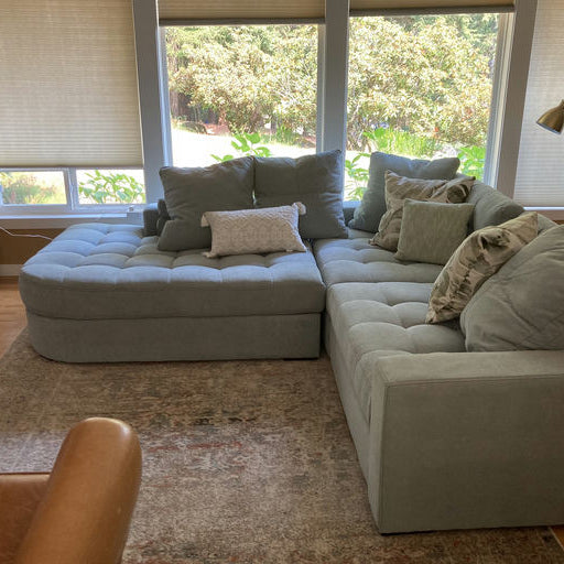 A cozy light blue sectional sofa with multiple pillows sits in a bright room, alongside a wooden cabinet and a fan near large windows.
