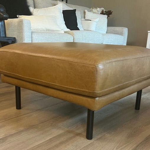 A stylish tan leather ottoman with black legs sits on wooden flooring, in a cozy living room featuring a light-colored sofa and decorative pillows.