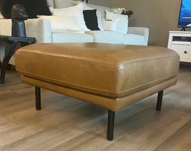 A stylish tan leather ottoman with black legs sits on wooden flooring, in a cozy living room featuring a light-colored sofa and decorative pillows.