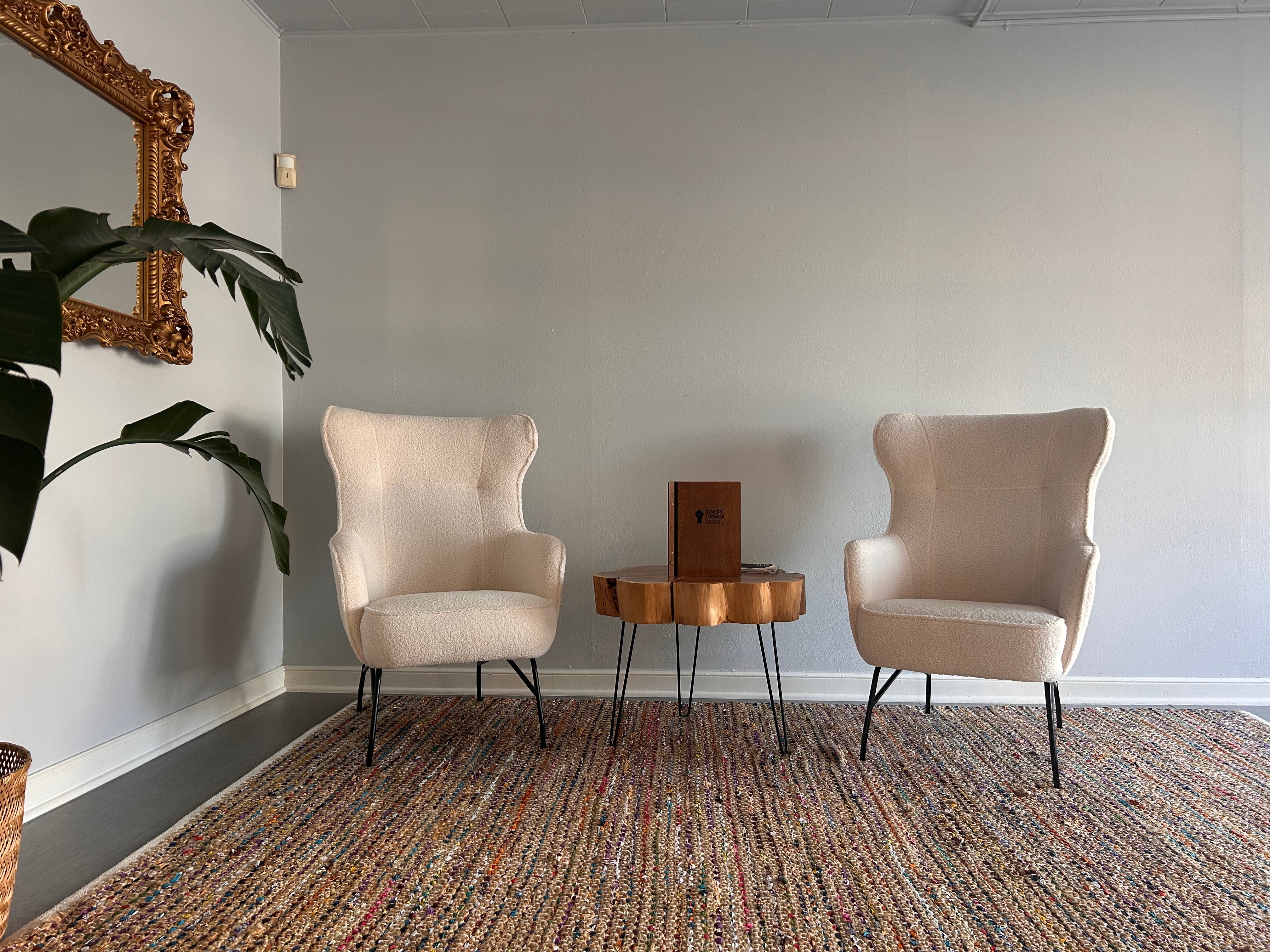 Cozy seating area featuring two cream-colored chairs, a wooden side table, and a decorative mirror, complemented by a textured area rug and plant.