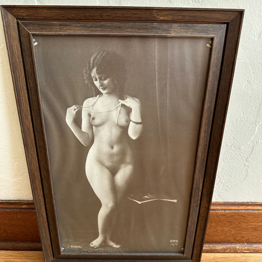 Framed sepia photograph of a woman in a classic pose, showcasing her figure, standing on one leg and holding a necklace, with a soft background.