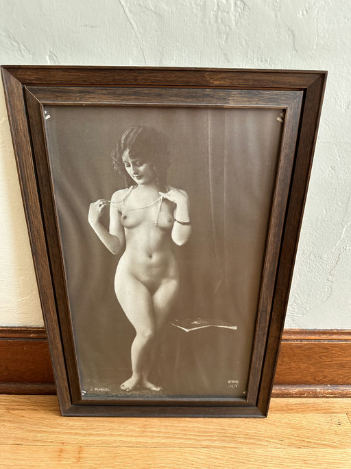 Framed sepia photograph of a woman in a classic pose, showcasing her figure, standing on one leg and holding a necklace, with a soft background.