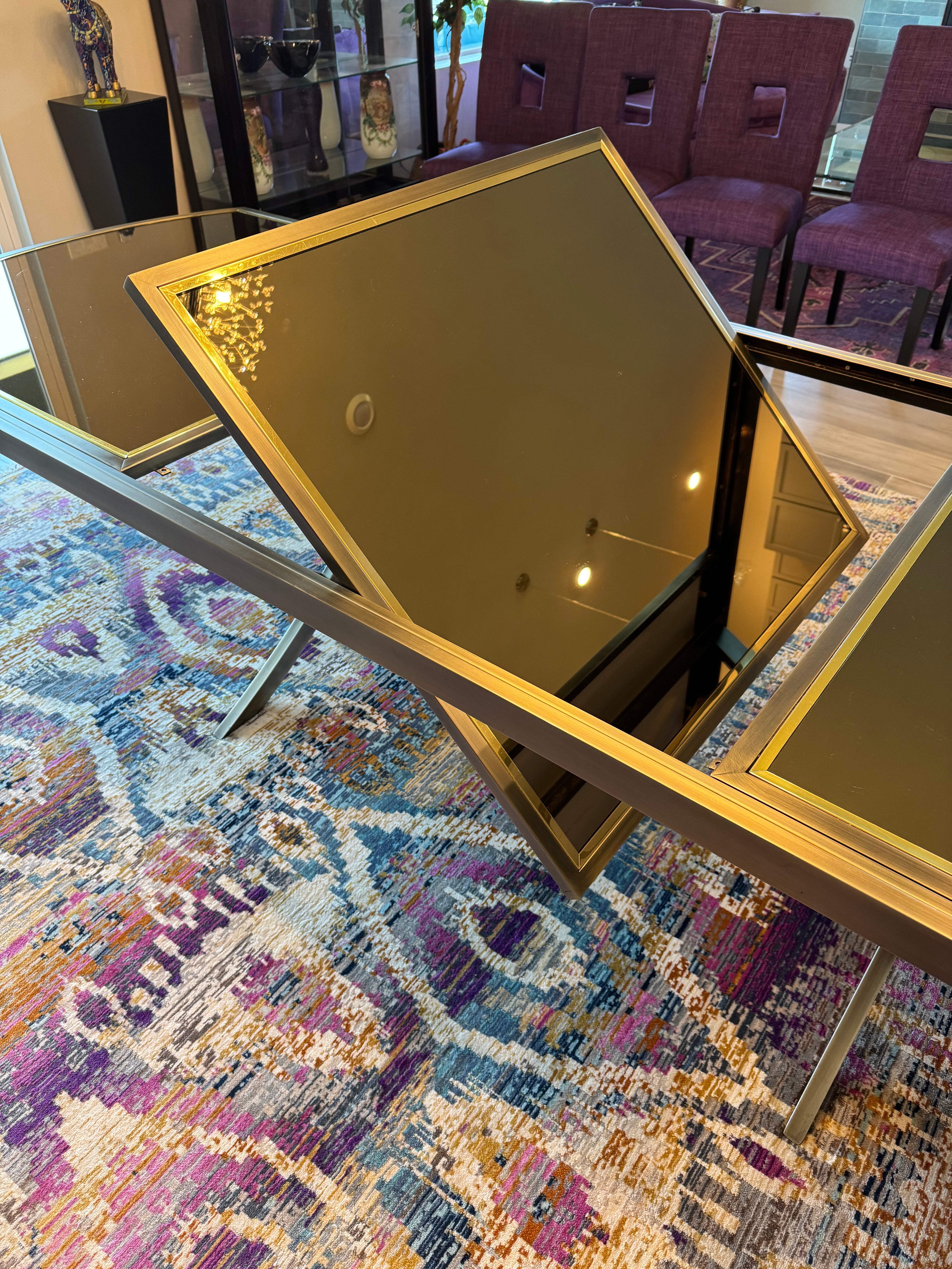 A sleek, modern coffee table with glass surfaces sits on a colorful patterned rug. Background features purple chairs and decorative items.