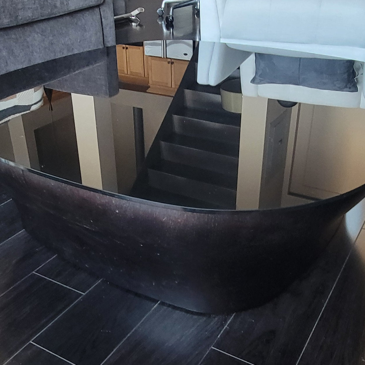 A modern, oval coffee table with a dark wooden base and reflective glass top is shown in a living room setting with a couch and staircase.