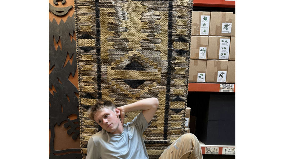 A young man poses against a patterned rug in a store, surrounded by boxes. The background features rustic decor and natural elements.