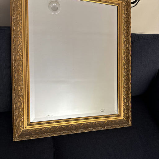 A vintage mirror with an ornate gold frame rests against a dark blue fabric sofa, reflecting light in a cozy indoor setting.