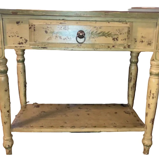 A vintage yellow side table with a single drawer, featuring worn paint and decorative detailing. It's positioned in front of a TV.