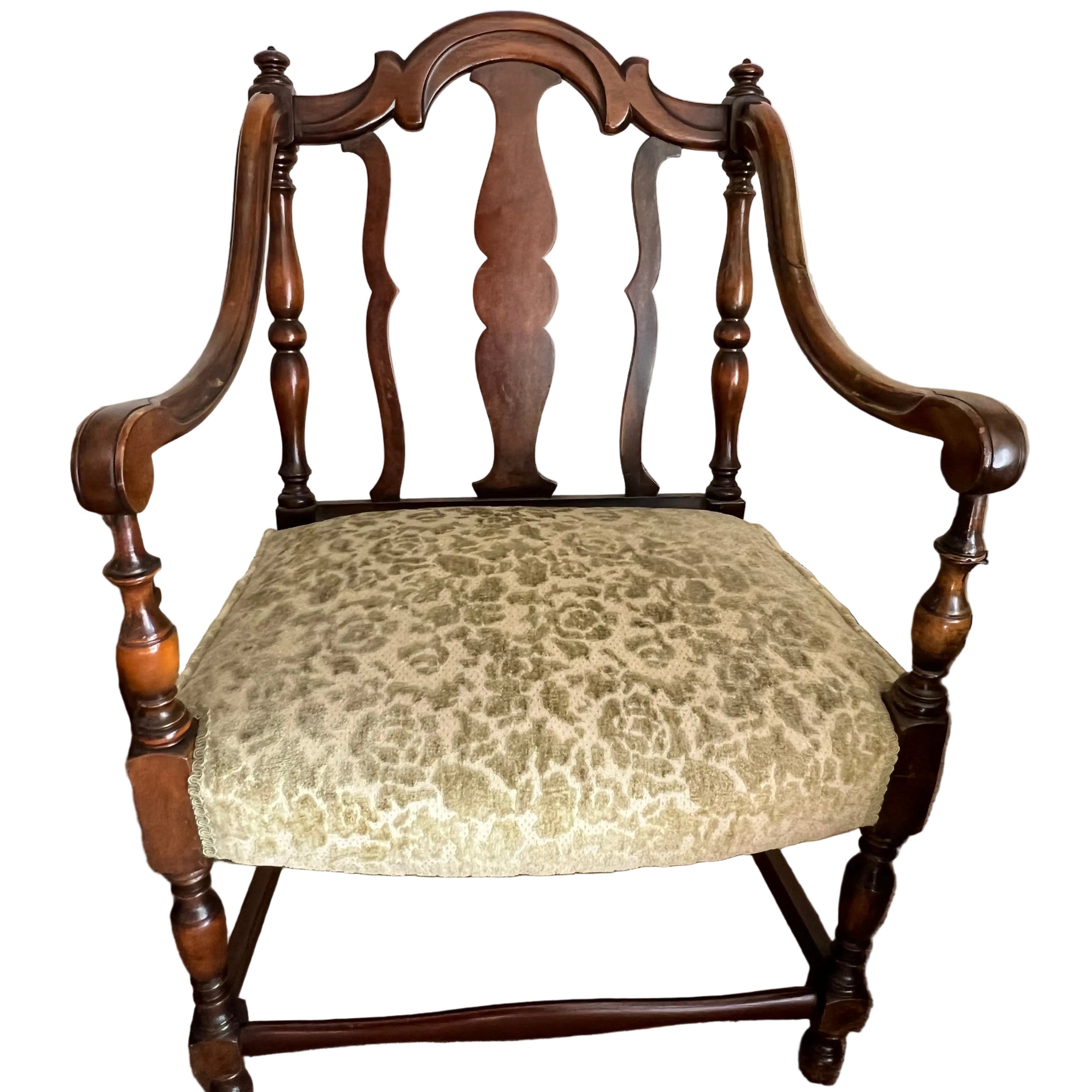 A vintage chair with a wooden frame, featuring an ornate backrest and green patterned upholstery. It rests on a carpeted floor beside a table.