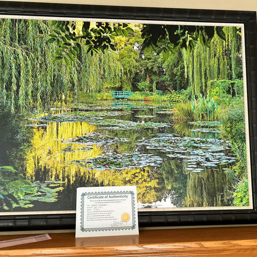 A vibrant landscape painting featuring a serene pond surrounded by lush greenery and water lilies, displayed alongside a certificate of authenticity.