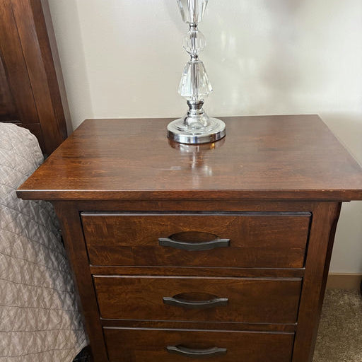 A wooden nightstand with three drawers, featuring black handles. A crystal lamp with a gray shade is placed on top, beside a bed with a quilted cover.