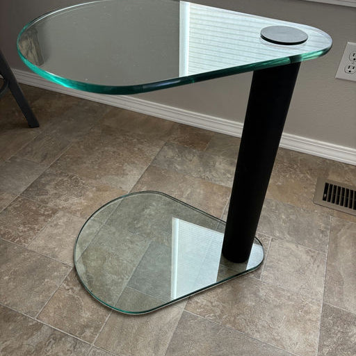 A modern, two-tier glass side table with a black cylindrical base. The top surface is oval-shaped, resting beside a window on a tiled floor.