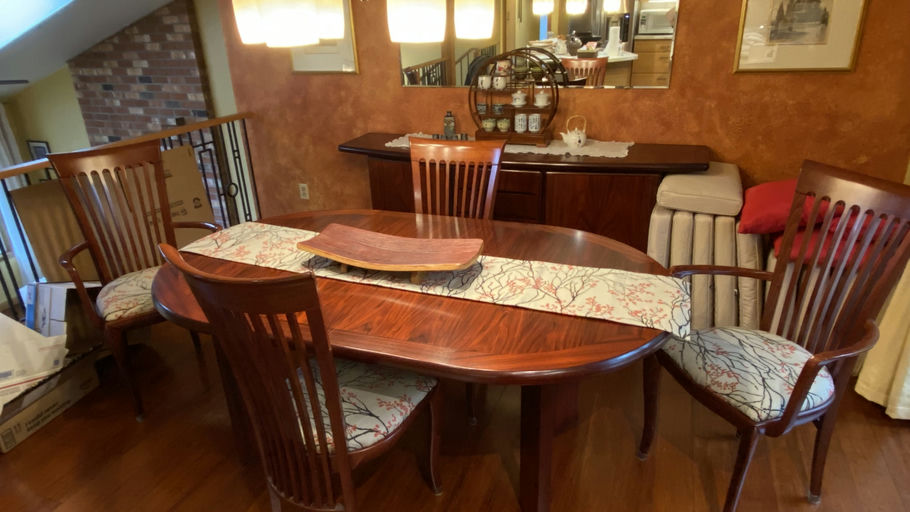 A cozy dining area featuring a wooden table with a decorative runner, surrounded by eight wooden chairs, and a hutch displaying teacups.