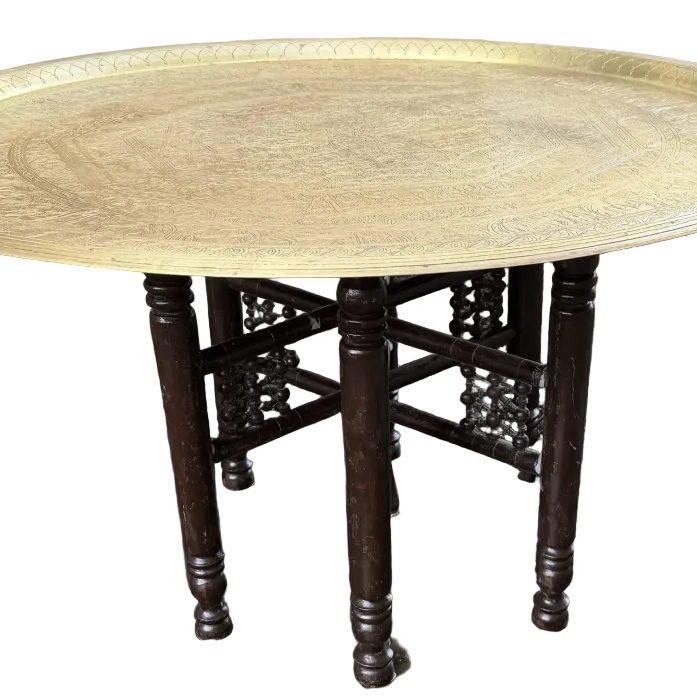 A round brass tray rests on a dark wooden table with four legs, set on a gravel surface next to a house with beige siding.