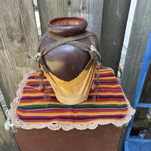 A rustic brown ceramic pot sits on a colorful striped textile with lace trim, adorned with wooden beads and leather straps against a wooden fence.