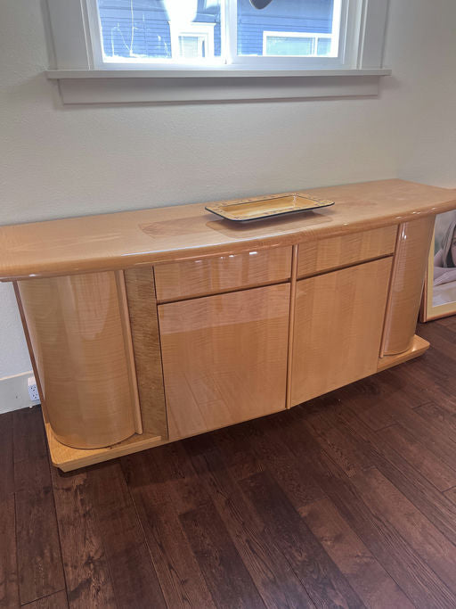 A polished wooden sideboard with rounded edges sits beneath a window, featuring two drawers and a decorative tray on top, on a wooden floor.