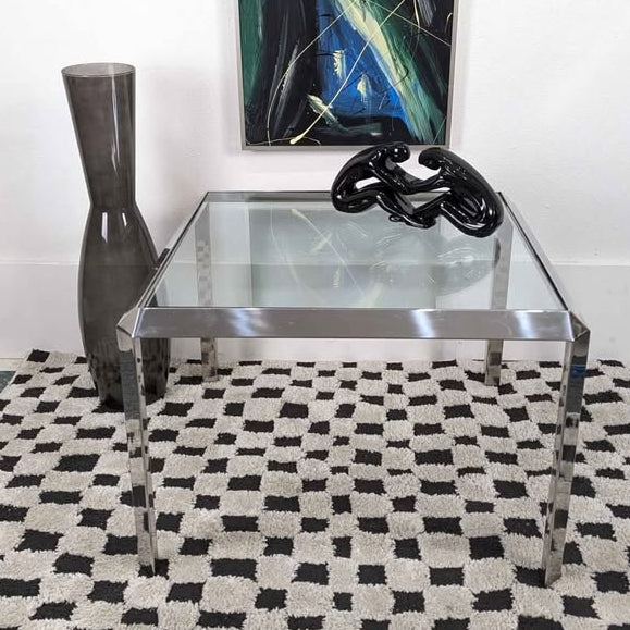 A glass-top coffee table with metal legs sits on a checkered rug. A tall gray vase and a black sculpture are displayed on the table beside a colorful painting.