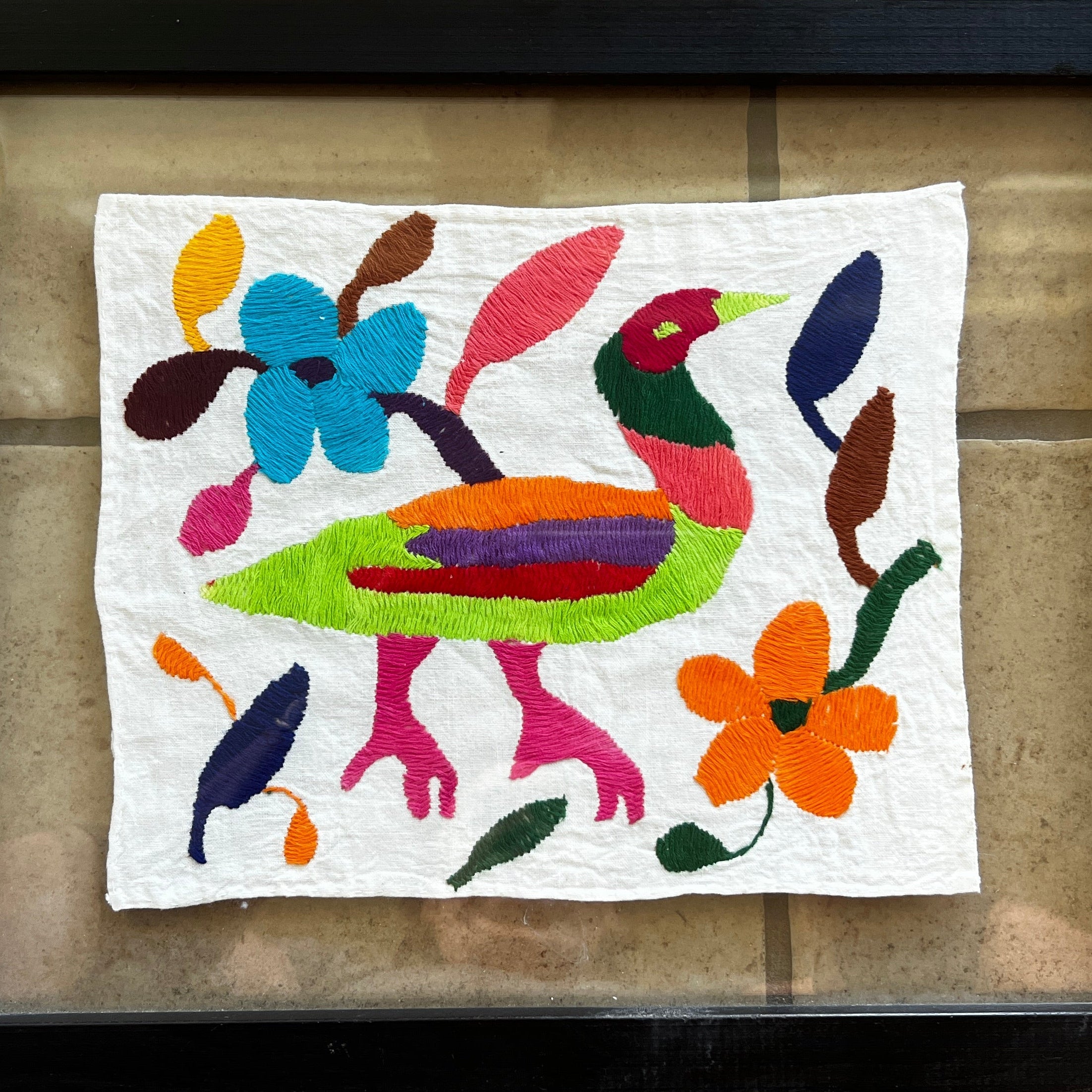A colorful embroidered artwork featuring a stylized bird surrounded by flowers and vibrant shapes, framed against a tiled background.
