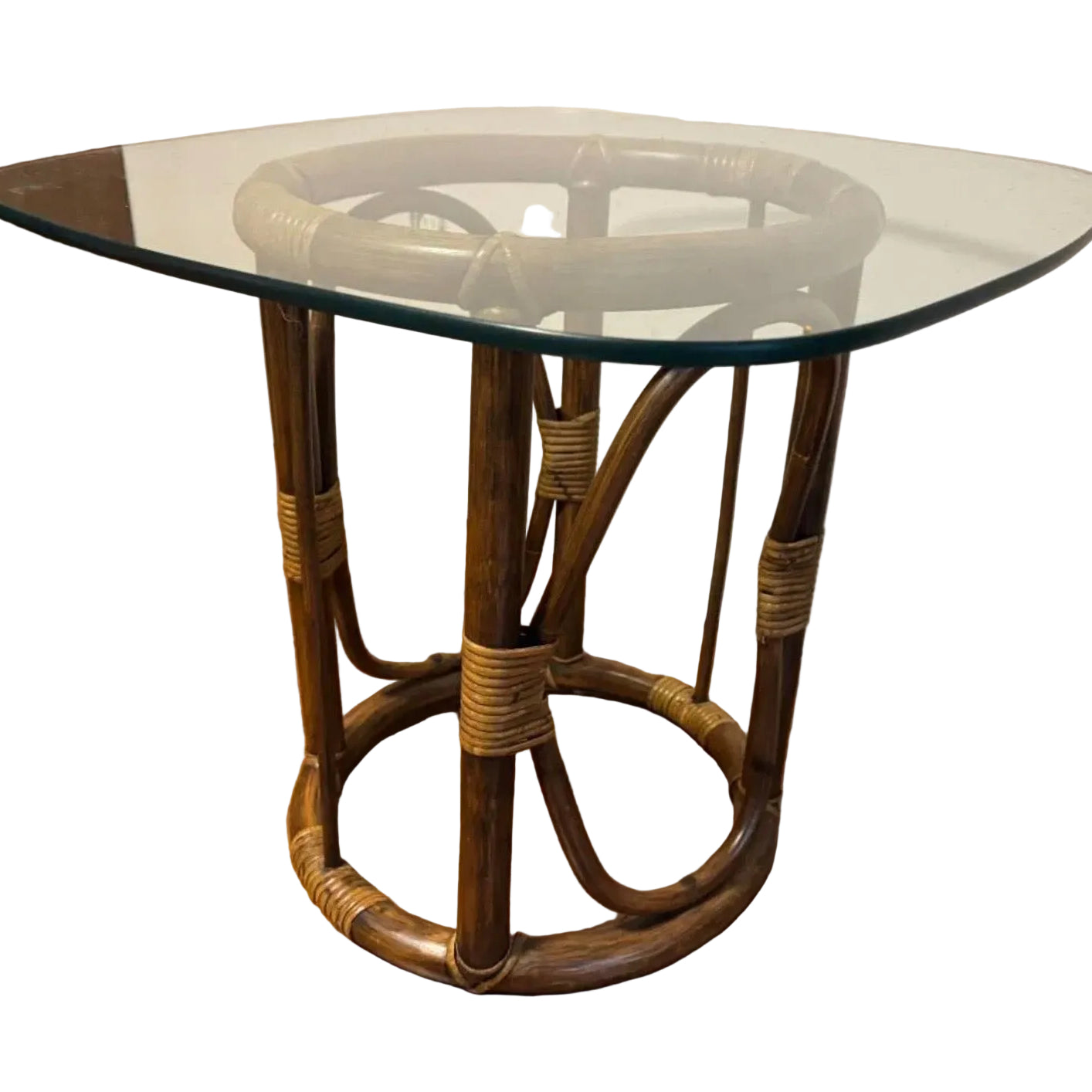 A small, round table with a glass top and a woven rattan base, set on a wooden floor with a wall behind it. Simple, modern design.