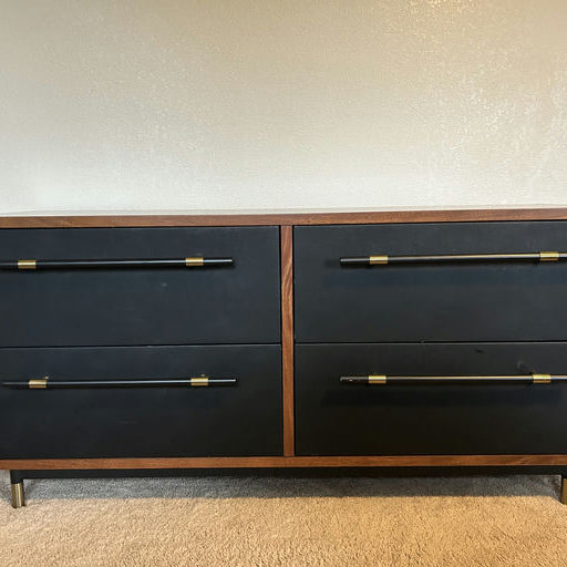 A modern dresser with a wooden frame, featuring four black drawers, each with sleek black handles accented by gold hardware. Set on metal legs.