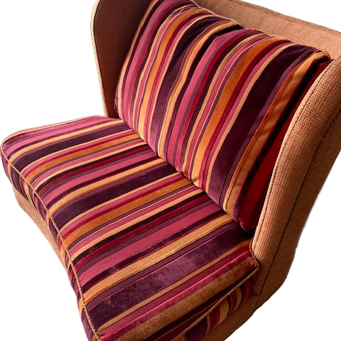 A striped loveseat with vibrant orange, pink, and purple upholstery sits on a gray carpet next to a cardboard box. Natural light filters in.