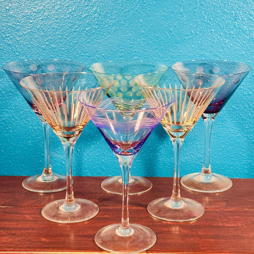 A collection of colorful, patterned martini glasses arranged in a semi-circle against a vibrant turquoise background, resting on a wooden surface.