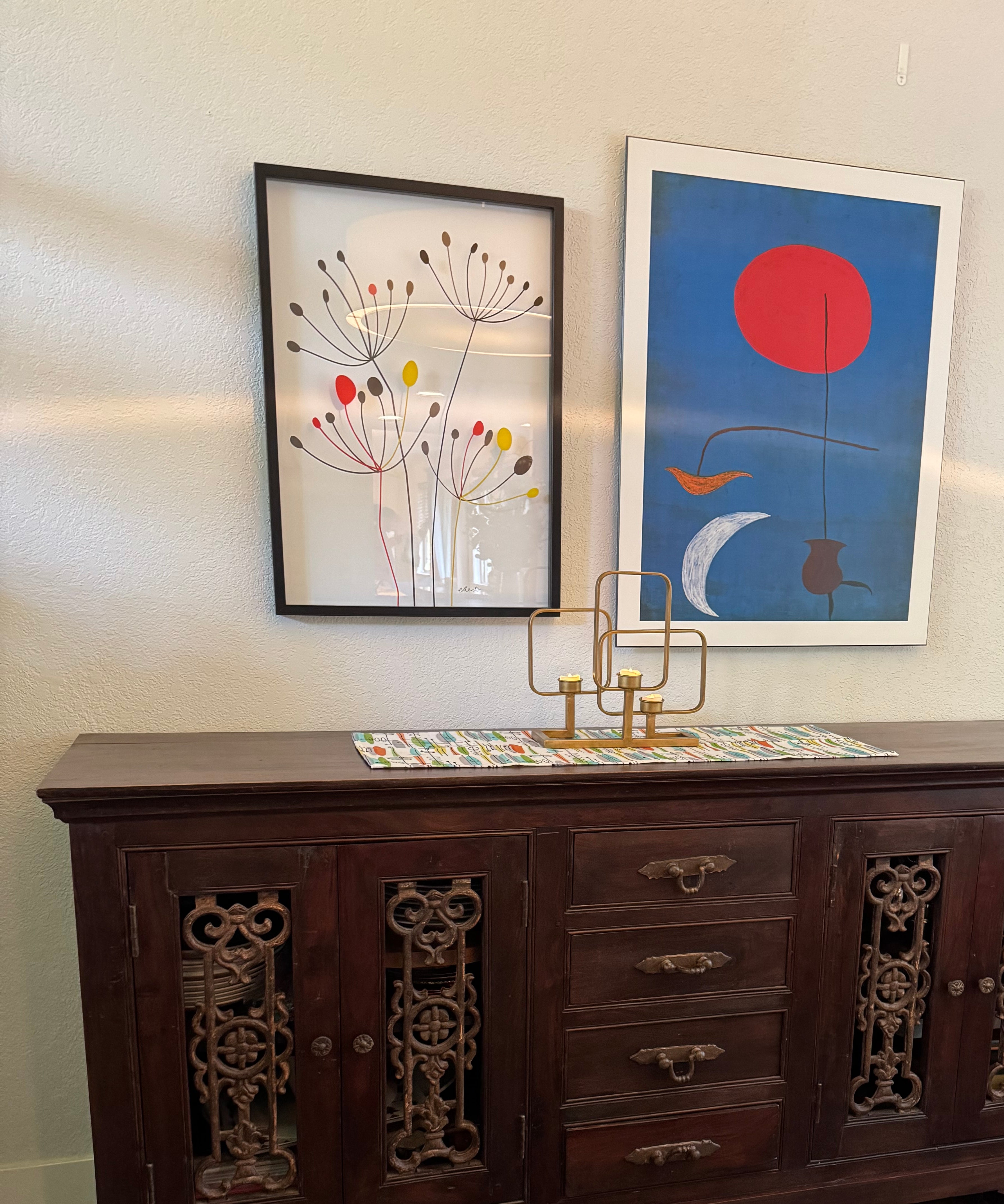 A wooden sideboard with intricate carvings, topped with a colorful table runner. Two framed artworks hang above it: one abstract, one floral.