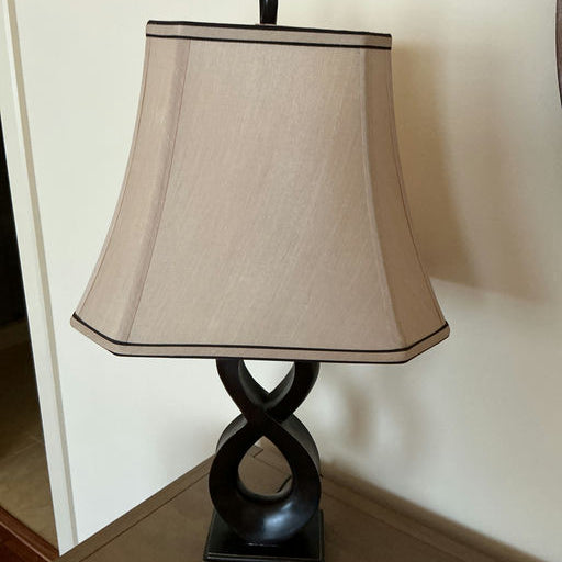 A brown lamp with an infinity-shaped base and a beige shade sits on a wooden table. The background features a plain wall.