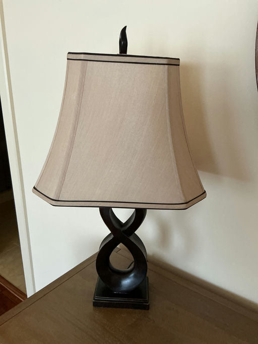 A brown lamp with an infinity-shaped base and a beige shade sits on a wooden table. The background features a plain wall.