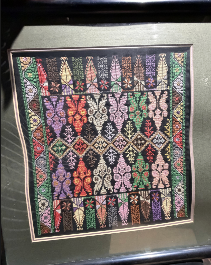 Colorful textile artwork features intricate geometric patterns and stylized motifs in vibrant hues, framed on a black background.