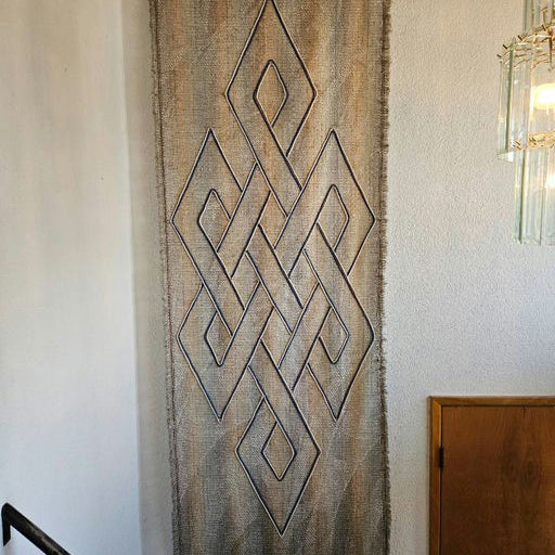 A woven wall hanging featuring an intricate geometric pattern in earthy tones, displayed against a light wall near a staircase and wooden door.