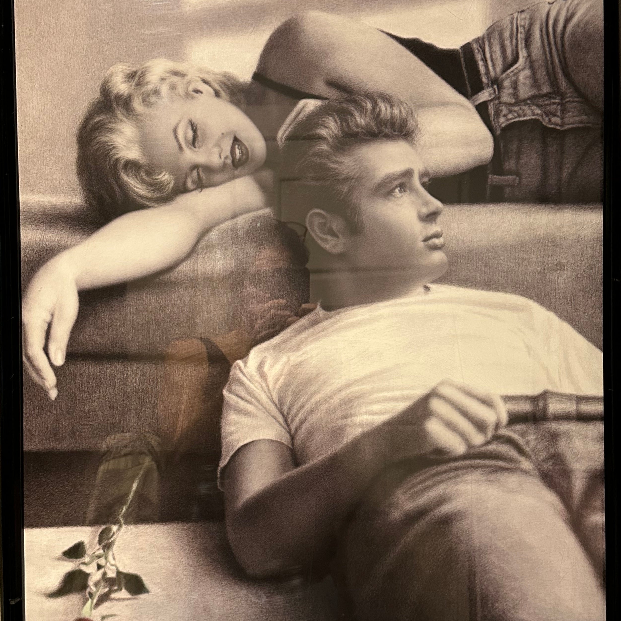 A black-and-white framed print features a smiling woman with blonde hair lounging beside a thoughtful man, a rose resting on the surface below.