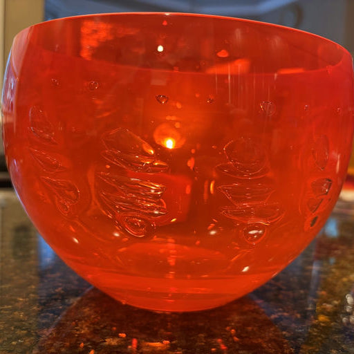 A bright red plastic bowl with decorative leaf patterns rests on a dark countertop, reflecting warm light and showcasing its glossy surface.