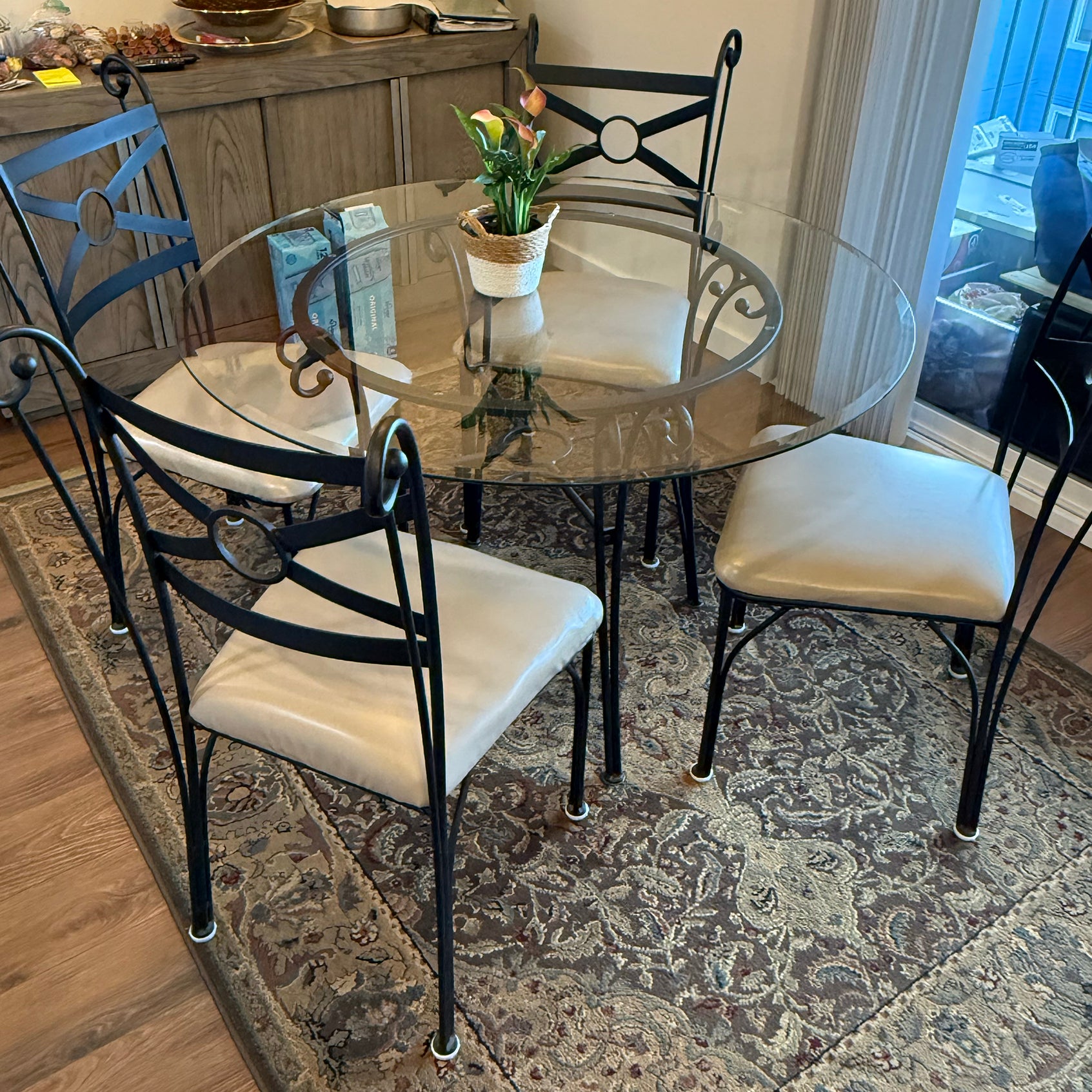 A round glass dining table with four ornate black metal chairs, set on a patterned rug. A small potted plant sits in the center, adding a touch of decor.