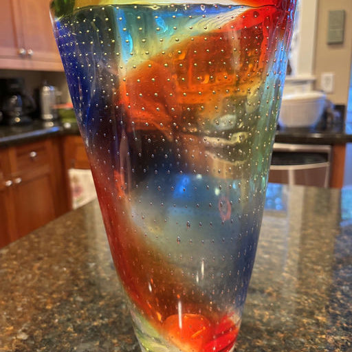 A colorful glass vase with swirling red, blue, green, and yellow hues, decorated with small bubbles, sitting on a dark granite counter.