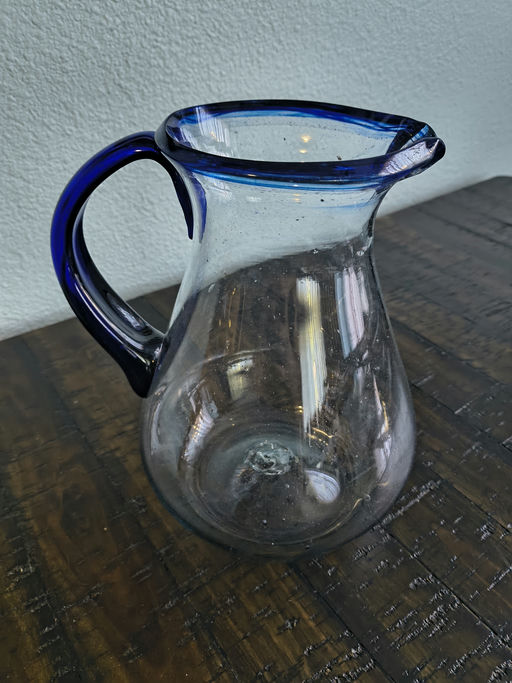 A clear glass pitcher with a blue rim and handle rests on a wooden table, showcasing its elegant design against a textured background.