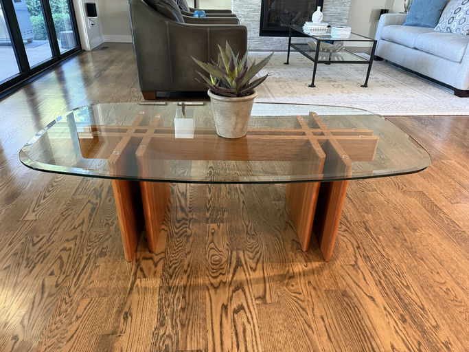A modern coffee table featuring a glass top and wooden base with a minimalist design. A potted plant adds a touch of greenery to the setting.