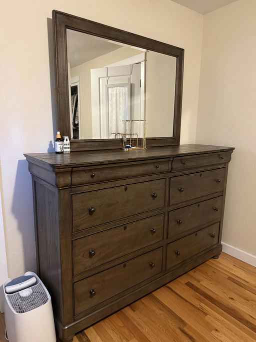 A wooden dresser with multiple drawers sits against a wall, topped with a large mirror. A small decorative house and a gold accessory holder are visible.