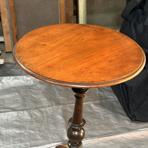 A wooden side table with a round top and a decorative, turned pedestal base, set against a backdrop of blurred furniture and a dark cloth.