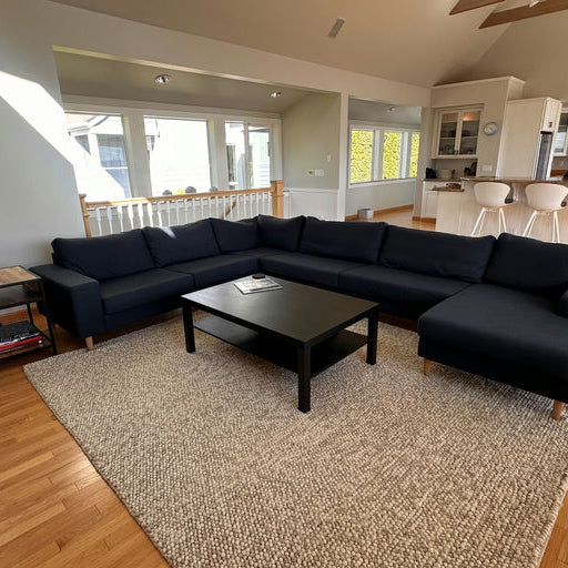 A cozy living room with a dark sectional sofa, black coffee table, and light wooden flooring. Sunlight streams in through large windows.