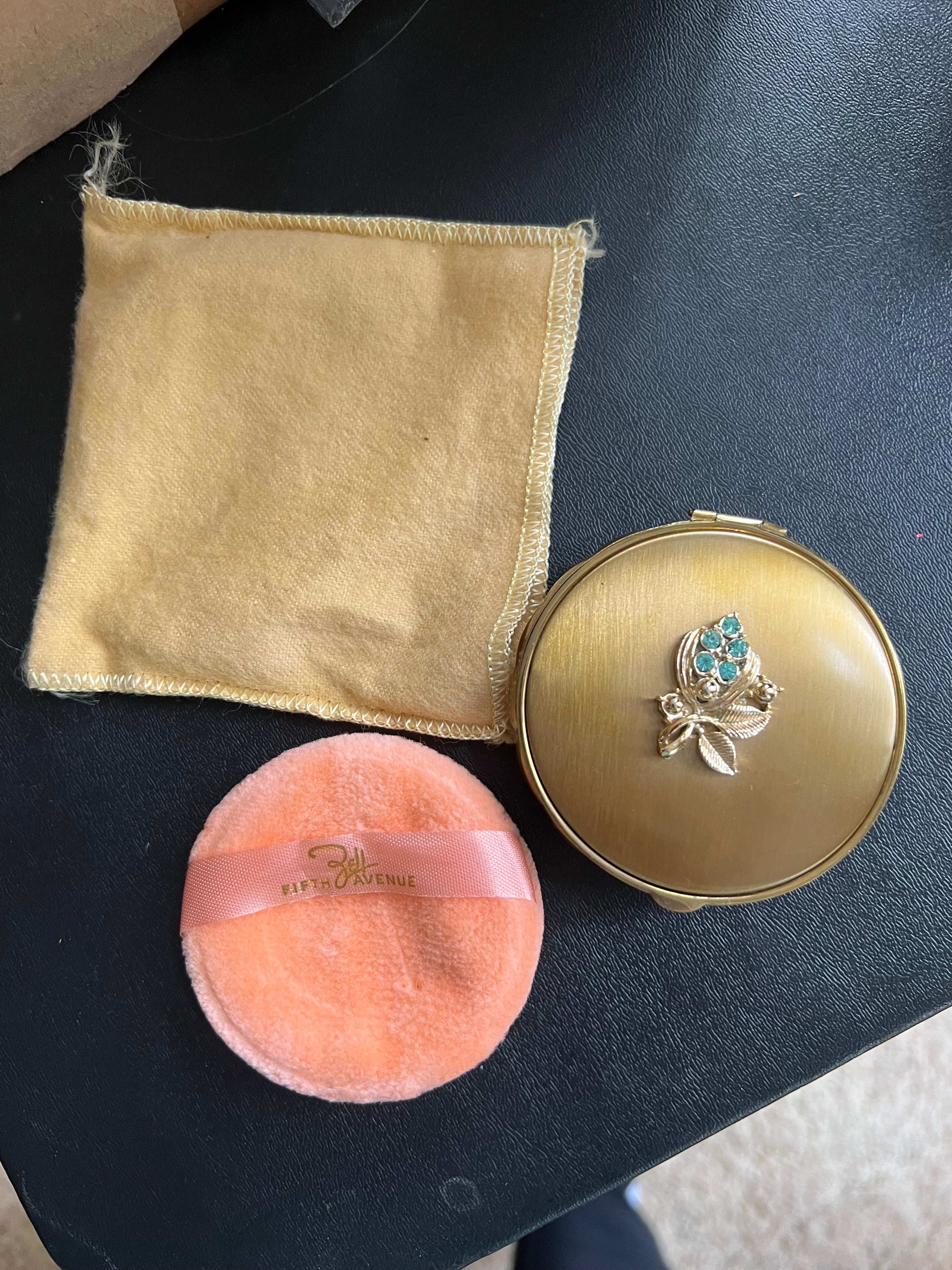 A gold compact mirror with a floral design, an orange puff, and a yellow cloth are arranged on a dark surface. The cloth shows stitching details.