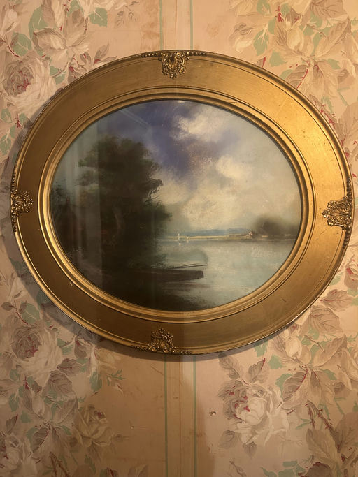 A framed landscape painting featuring a serene water scene with trees, a boat, and a cloudy sky, set against floral wallpaper.